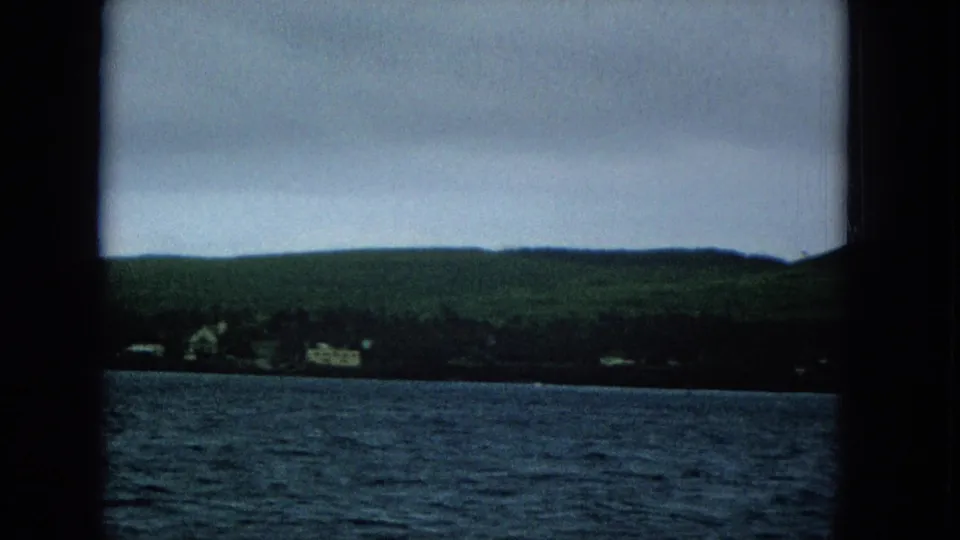 Final Super 8 frame looking from a boat toward Molokai shoreline buildings, green hills, and choppy water beneath a gray sky.