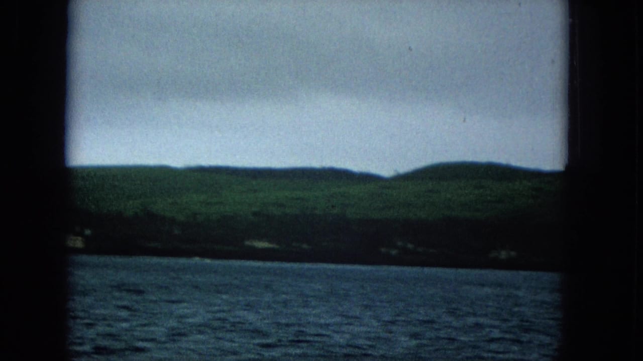 1960s home-movie view of a Molokai or Hawaii coastline from the water, with rolling green hills, scattered shoreline structures, and an overcast sky.