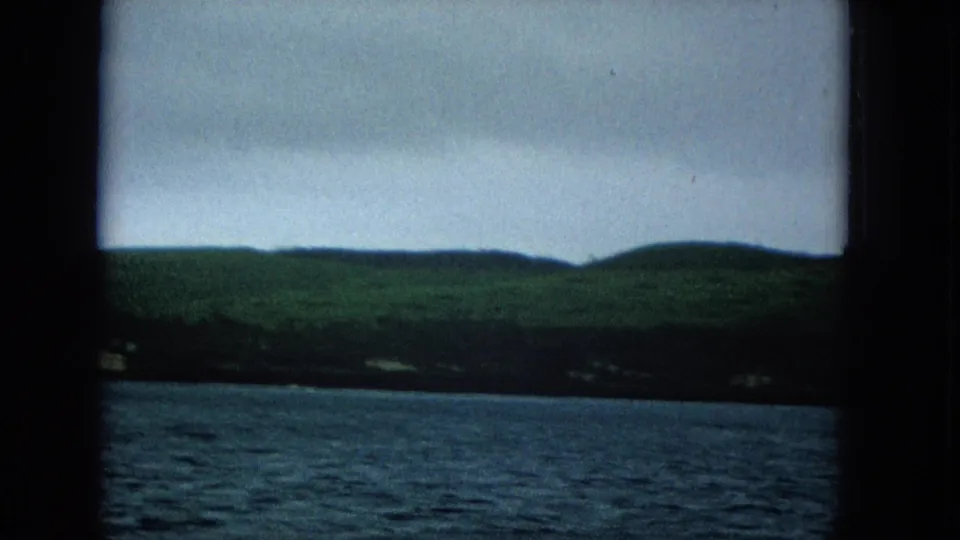 1960s home-movie view of a Molokai or Hawaii coastline from the water, with rolling green hills, scattered shoreline structures, and an overcast sky.