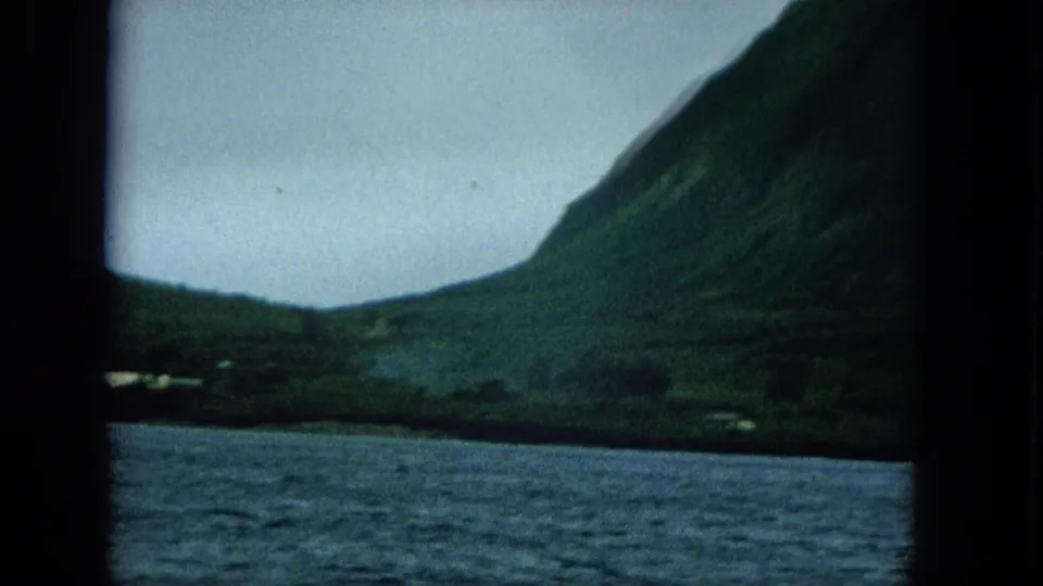 1960s home-movie frame from the water showing a steep green coastal slope, low shoreline settlement, and muted overcast light.