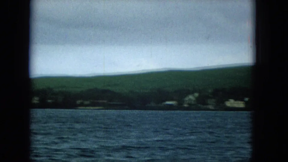 1960s boat-shot view of a low Molokai or Hawaii shoreline with blue water, scattered homes, open green hills, and heavy cloud cover.