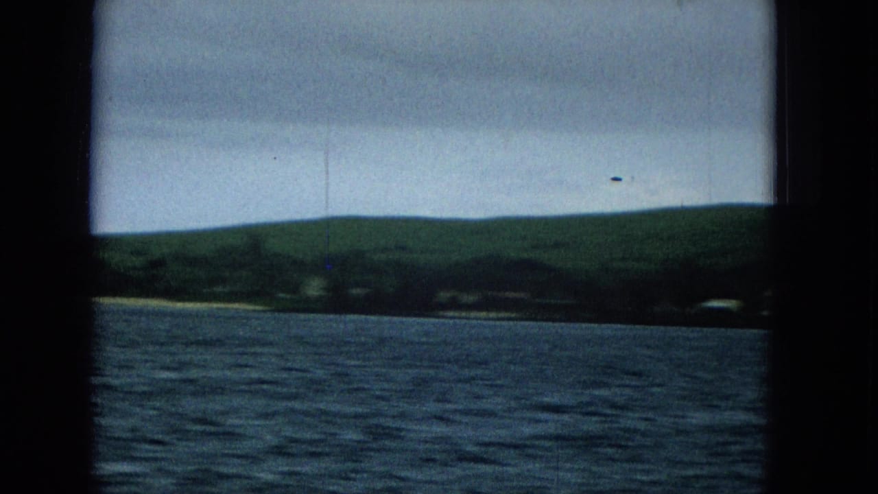 1965 home-movie frame showing rippled water in the foreground, a low green Molokai shoreline in the distance, and an overcast sky within dark 8mm frame borders.