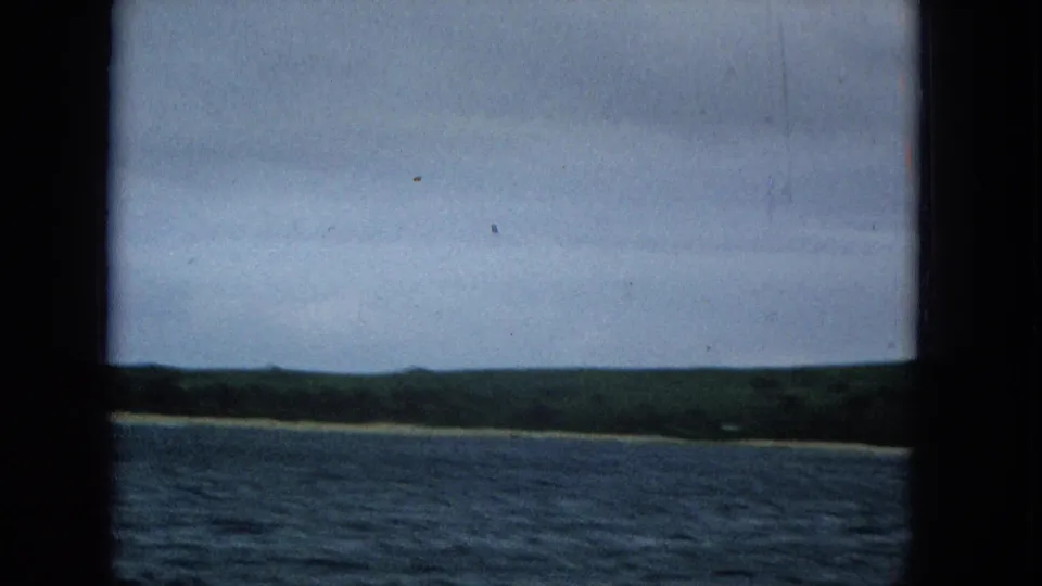 1965 home-movie frame looking across blue-gray water toward a low green shoreline, with dark vertical frame borders and muted color from the original small-gauge film.
