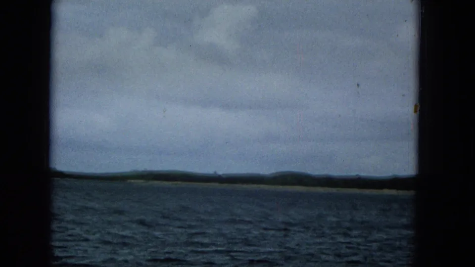 1965 home-movie frame with choppy water below a broad cloudy sky and a dark, low Molokai coastline stretching along the horizon.