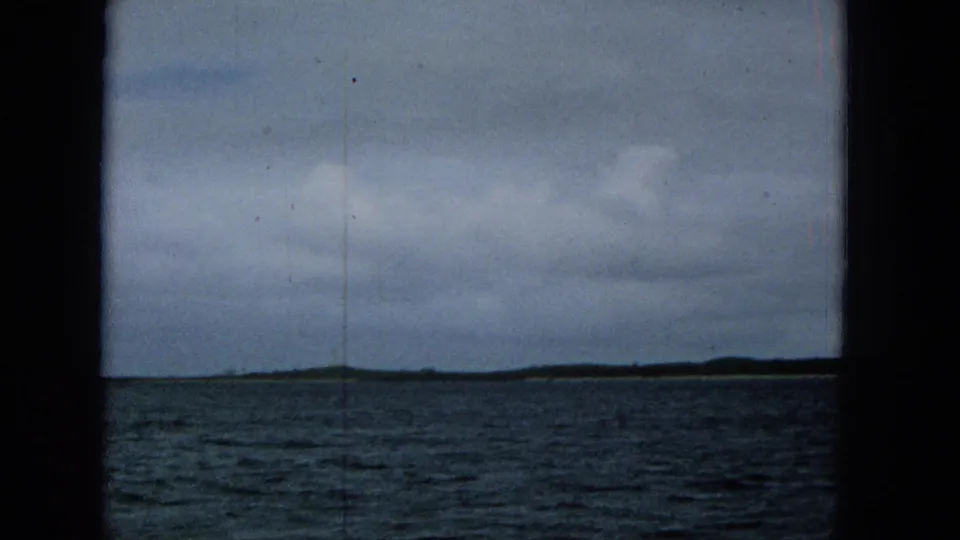 1965 home-movie frame showing rippled water, a low distant shoreline, heavy cloud cover, and a slender tower or vertical structure near the horizon.
