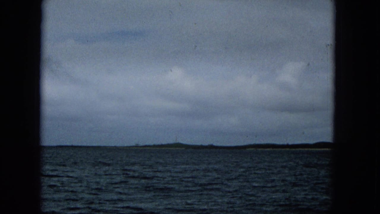 1965 Super 8 frame showing choppy ocean and a distant low island shoreline beneath a cloudy sky, filmed from a boat.