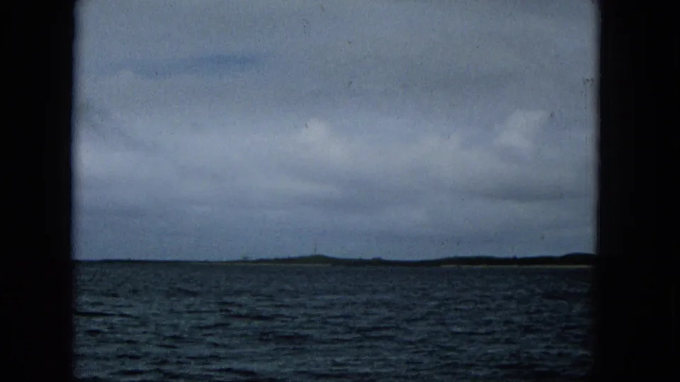1965 Super 8 frame showing choppy ocean and a distant low island shoreline beneath a cloudy sky, filmed from a boat.