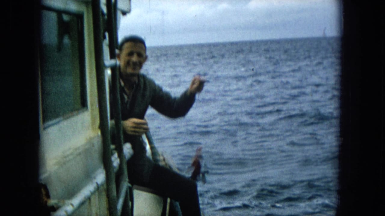 Smiling man on a boat holds up a small fish beside the open ocean.