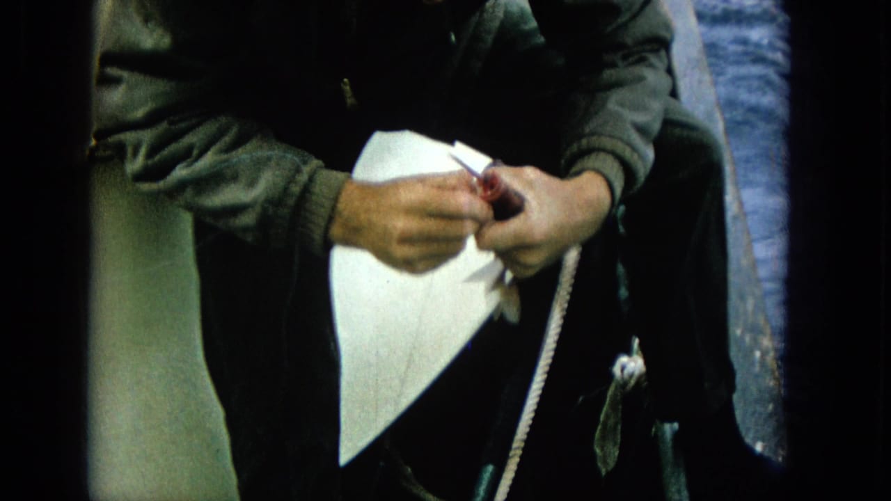 1965 Super 8 frame of an adult man in dark clothing on a boat, hands close to a fishing line or hook with blue ocean visible behind him.