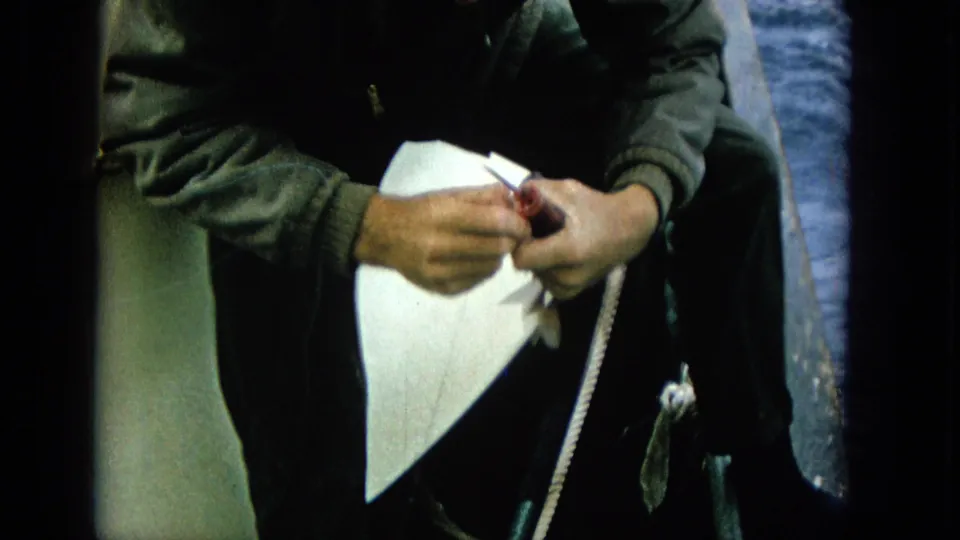 1965 Super 8 frame of an adult man in dark clothing on a boat, hands close to a fishing line or hook with blue ocean visible behind him.