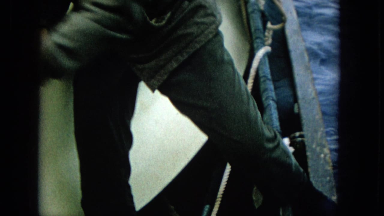 Adult man in dark clothing leans over the boat side in 1965 Super 8 footage, with ocean water and a white boat surface visible around him.