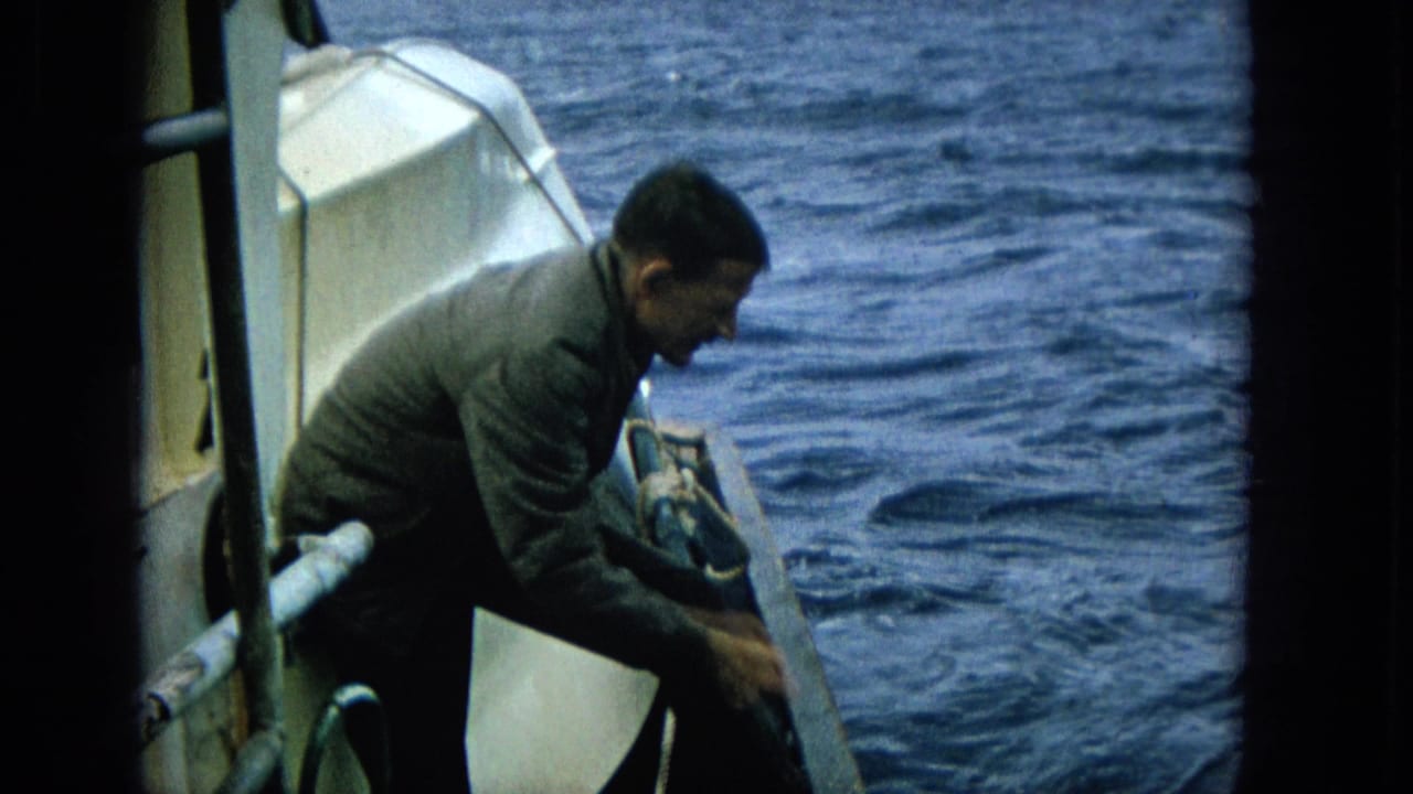 1965 Super 8 frame showing an adult man crouched at the edge of a boat, working near the rail with blue ocean filling the background.