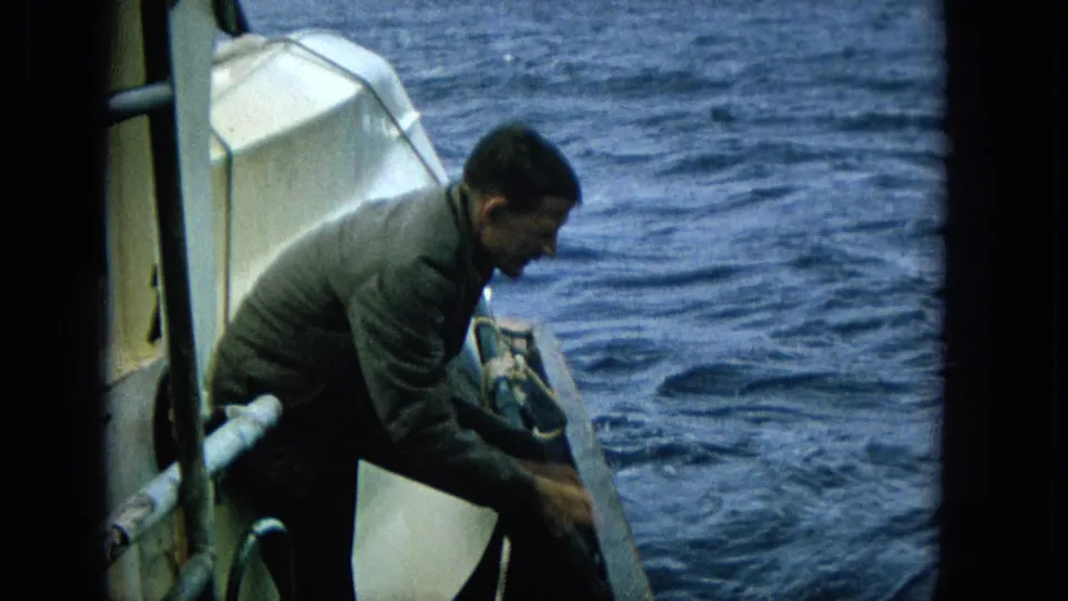 1965 Super 8 frame showing an adult man crouched at the edge of a boat, working near the rail with blue ocean filling the background.