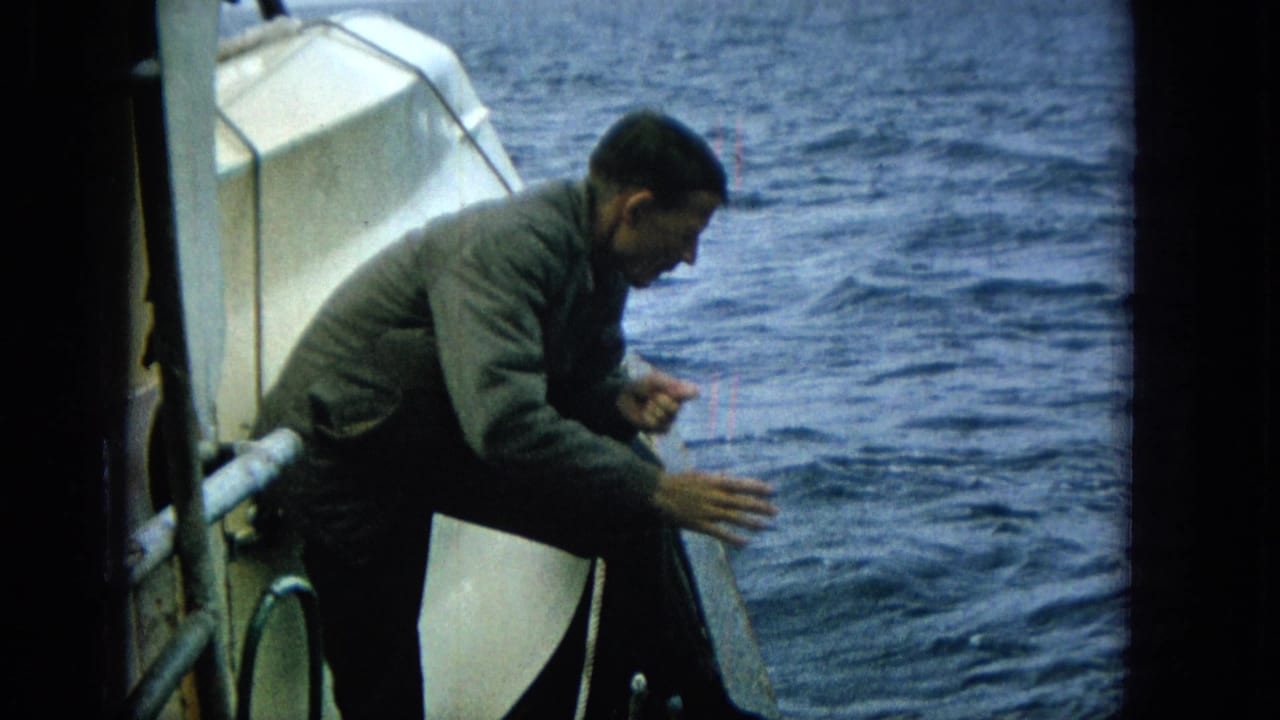 Adult man on a boat reaches outward over the ocean in 1965 Super 8 footage, appearing to handle a fishing line or catch at the rail.