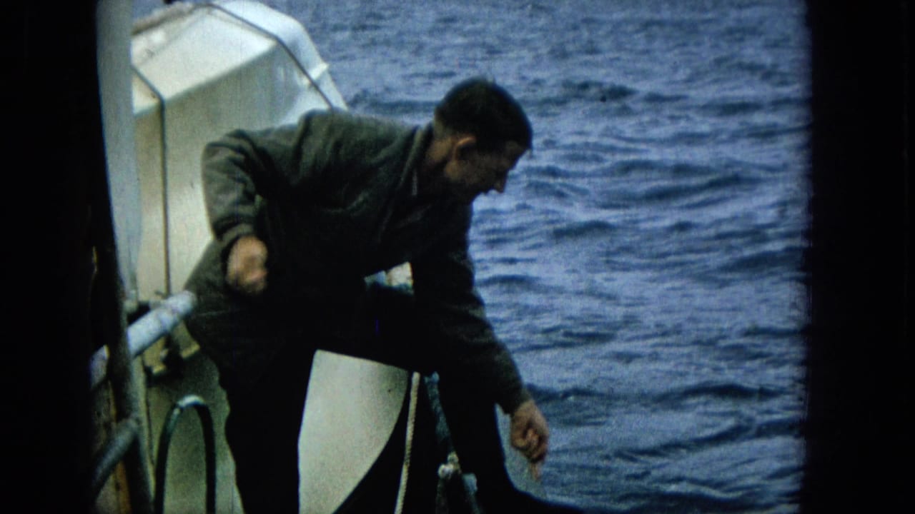 In 1965 home-movie footage, an adult man in a dark jacket leans over the side of a small boat while looking down toward the sea.