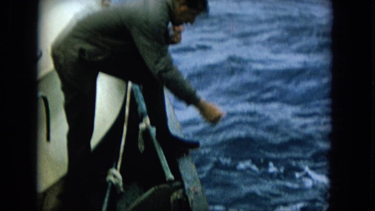 An adult man bends over the edge of a small boat, reaching down toward choppy open water during a fishing moment.