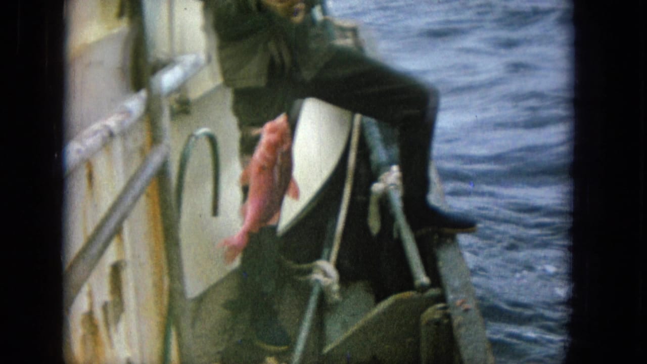 An adult man in dark clothing pulls a bright red fish up beside the small boat with open sea behind him.