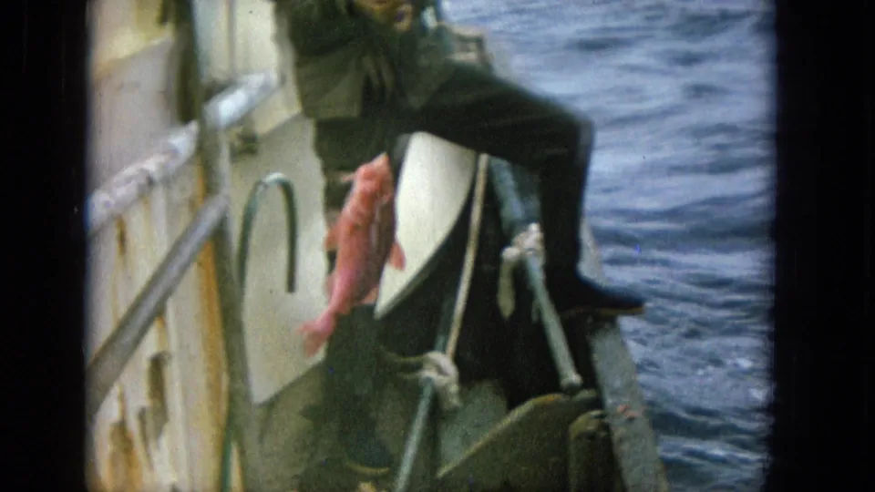 An adult man in dark clothing pulls a bright red fish up beside the small boat with open sea behind him.