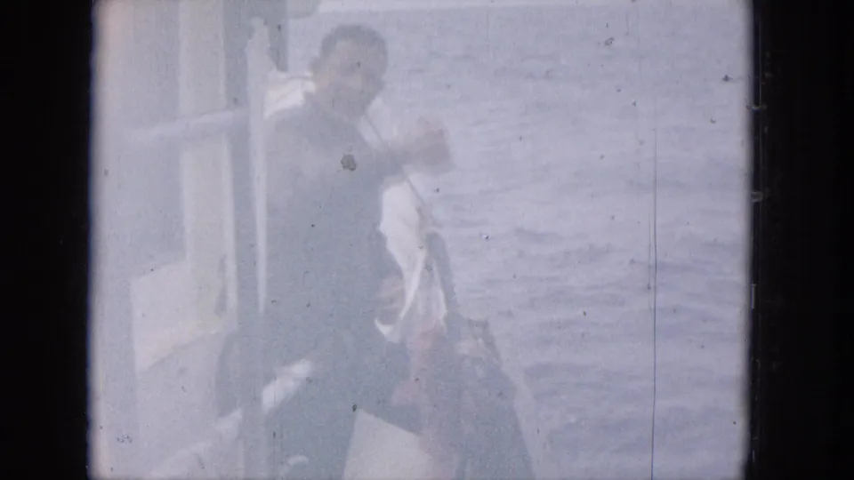 A soft, overexposed home-movie frame shows a man on the edge of a small boat with water in the background, his figure partly washed out by exposure and film fading.