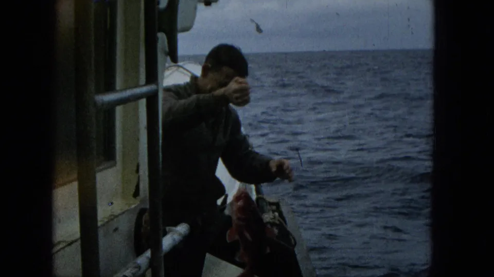 The man leans out from a small boat and holds a freshly caught fish near the rail, with open ocean and boat hardware visible in the background.