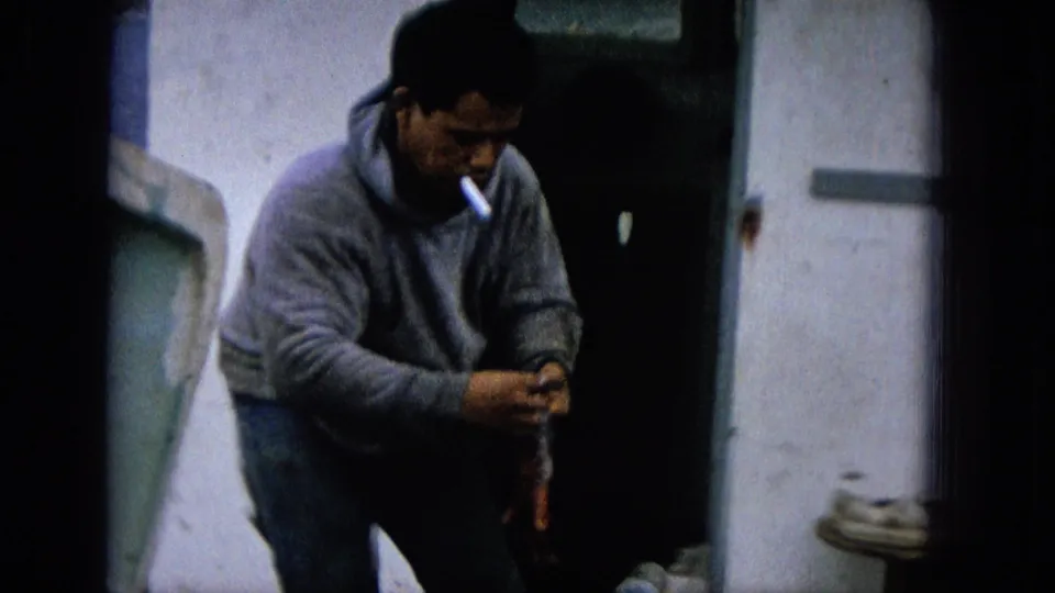 1965 8mm Molokai frame of an adult fisherman in a gray hooded sweatshirt smoking a cigarette while working with a fish beside a pale boat or dock structure.
