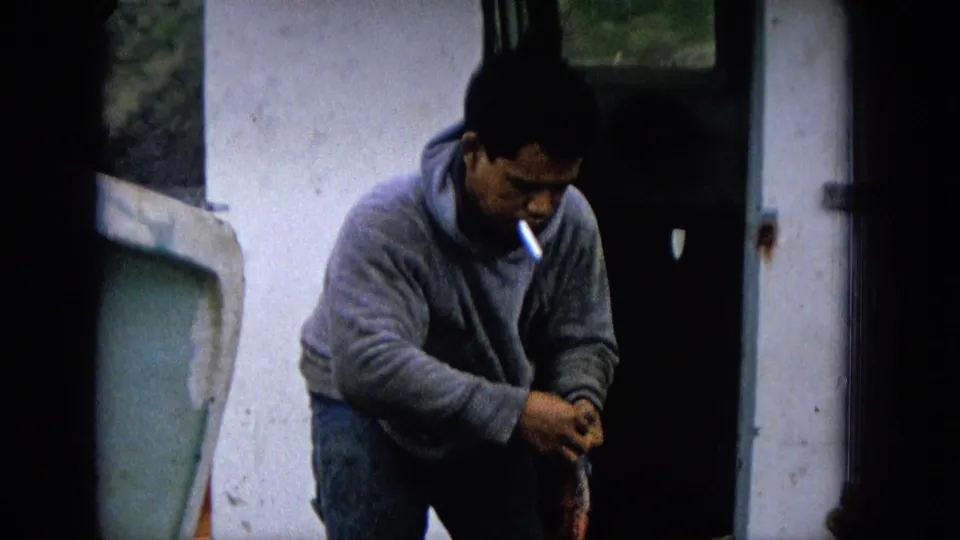 1965 8mm Molokai footage of an adult fisherman in casual clothes leaning over his catch, with a cigarette visible and a white boat or dock surface behind him.