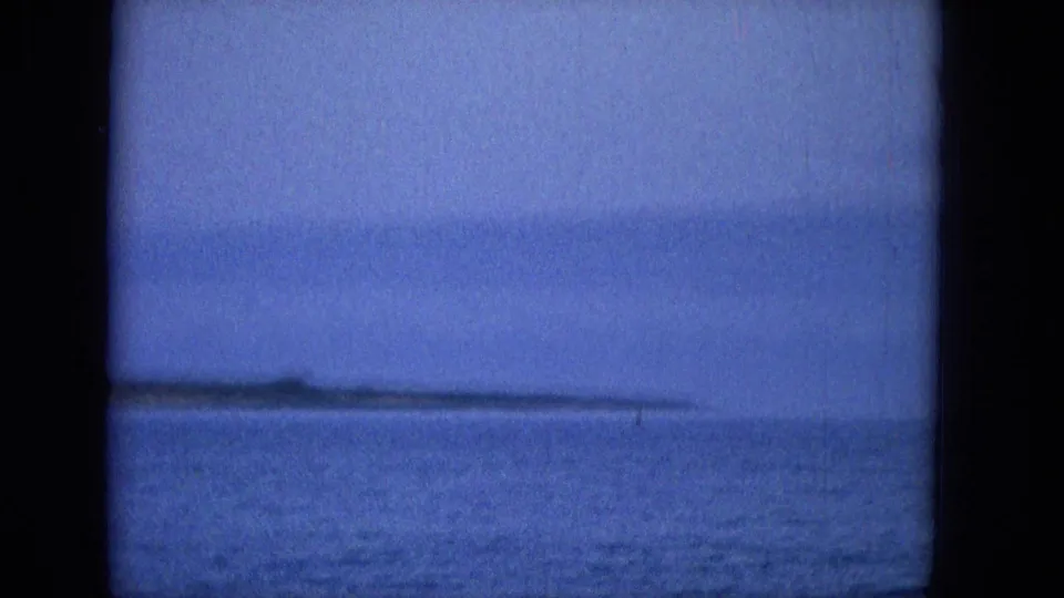 Blue dusk ocean view from the same 1965 8mm Molokai clip, with calm water and a distant island or shoreline silhouette on the horizon.