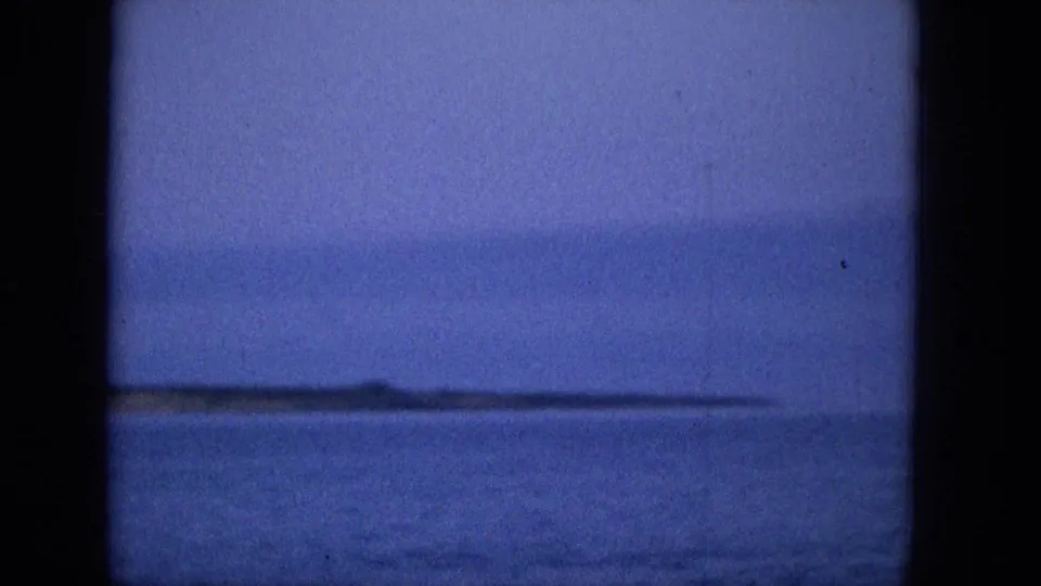 A blue-tinted 1965 home-movie frame shows open water and a distant low coastline viewed through dark vertical bars, consistent with a vessel or window perspective.