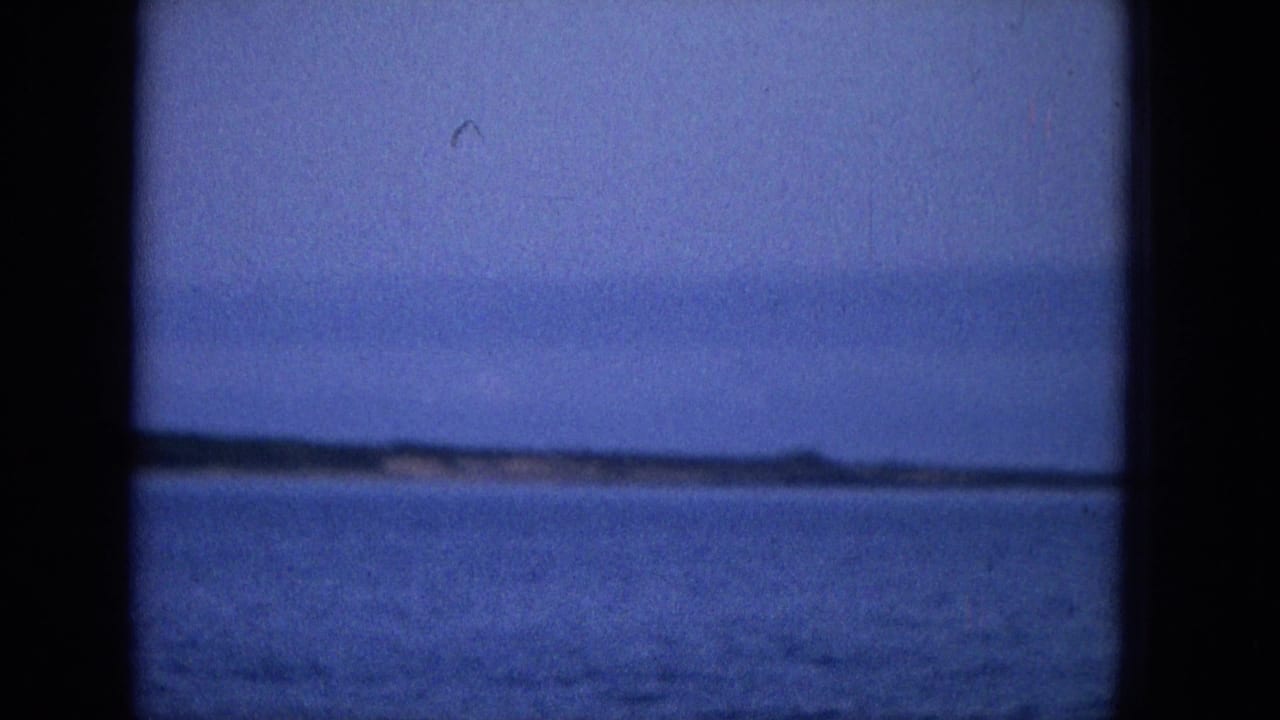 A distant shoreline and low landform appear across calm ocean water in a blue-tinted 1965 Hawaii travel-film frame.