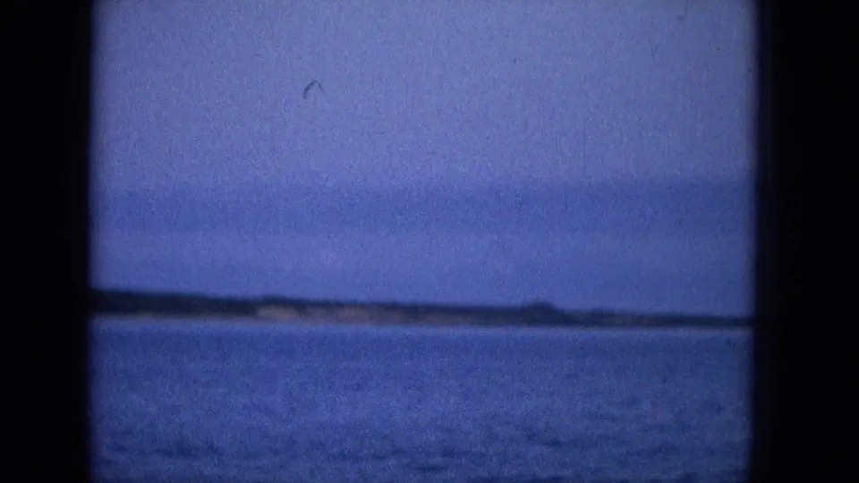 A distant shoreline and low landform appear across calm ocean water in a blue-tinted 1965 Hawaii travel-film frame.