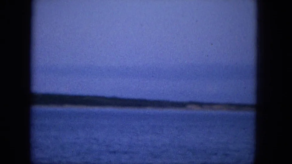 The frame shows open water, a low distant shore, and overcast sky through dark vertical framing, matching a passenger-shot coastal view from 1965 Hawaii footage.