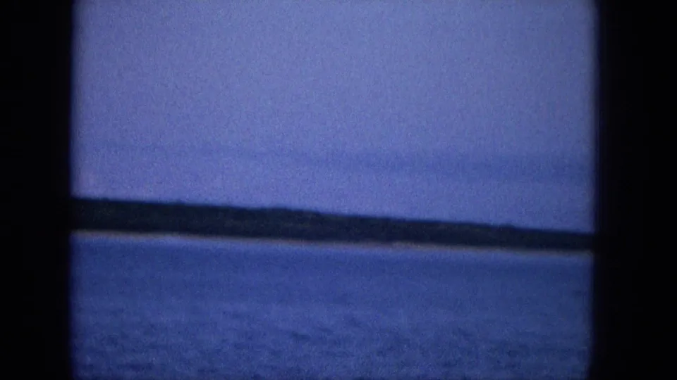 A sloping low coastline lies beyond blue ocean water, with dark frame edges and aged-film softness visible in this 1965 Hawaii coastal view.