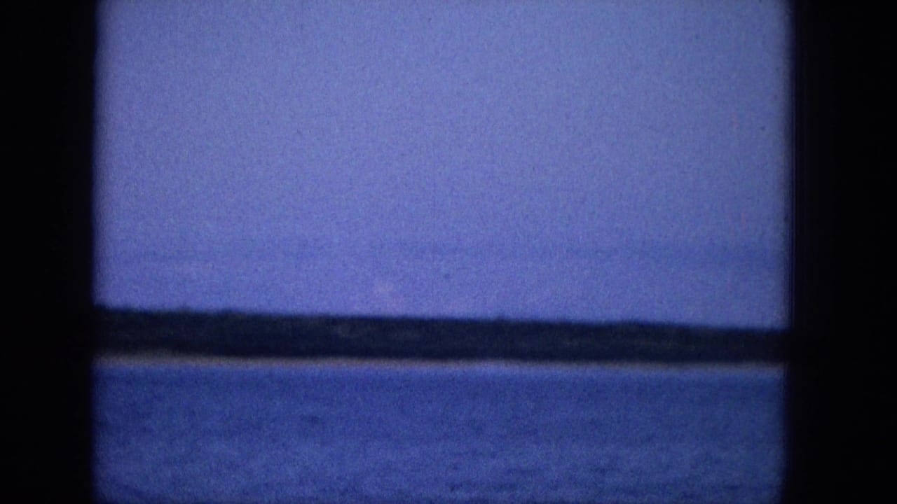 1965 Super 8 travel footage shows a cool-blue view of calm water and a faint shoreline or horizon seen through dark boat or window framing.