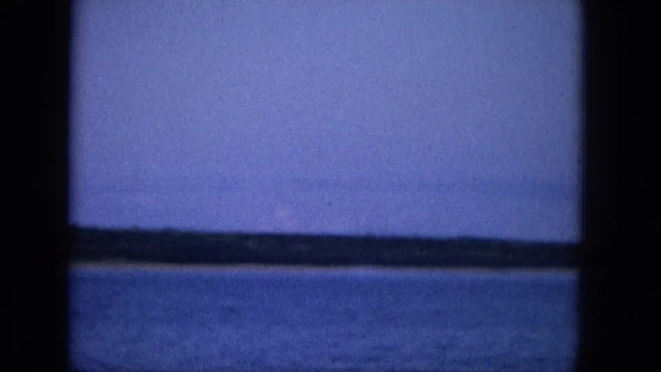 1965 Super 8 travel footage shows a cool-blue view of calm water and a faint shoreline or horizon seen through dark boat or window framing.