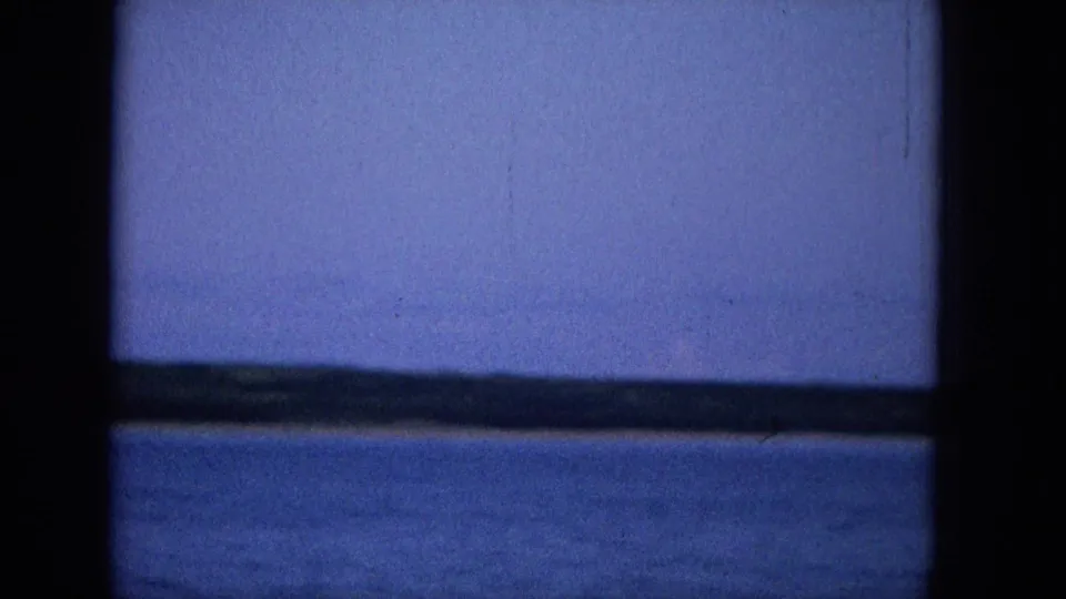 A cool-toned Super 8 frame records open water and a low distant horizon from behind a dark central frame or pole.