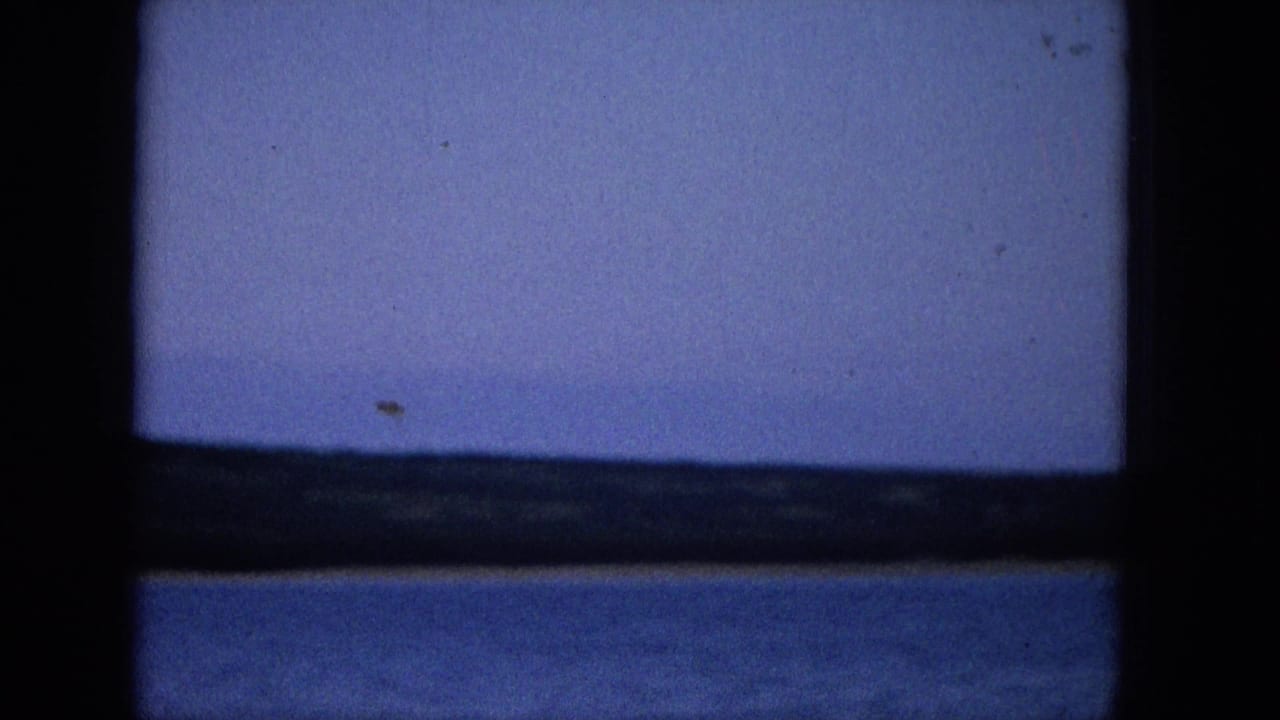 1965 Super 8 frame showing a coastal or lakeside view through a dark window frame, with blue water, distant low shore, and pale purple-blue sky.