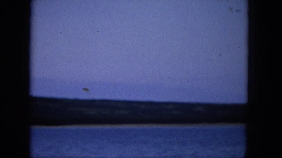 1965 Super 8 frame showing a coastal or lakeside view through a dark window frame, with blue water, distant low shore, and pale purple-blue sky.