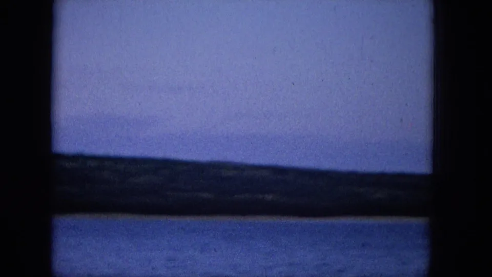 1965 Super 8 frame with a static view across blue water toward a low distant shoreline, divided by dark window or screen bars.