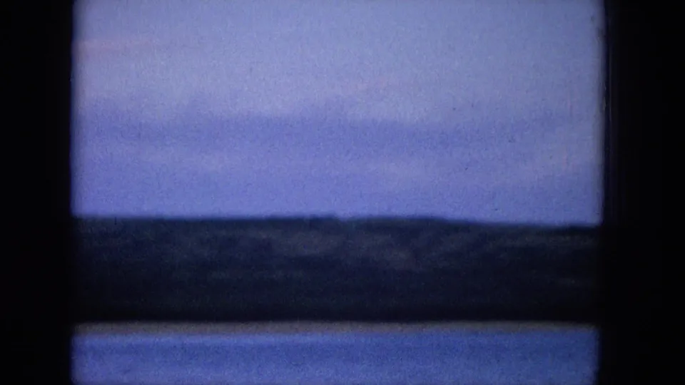 1965 Super 8 frame of blue water and a distant low shoreline seen through dark window panes, with subdued clouds and visible home-movie grain.