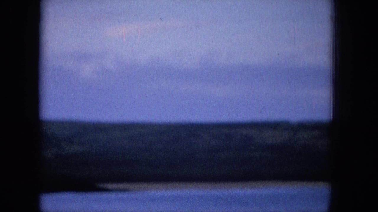 1965 archival home-movie frame showing a blue sky and dark coastal horizon viewed through a heavy window-like frame, with soft grain and low-light exposure.