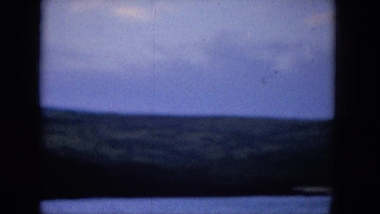 1965 archival home-movie view of a distant shoreline beneath a blue clouded sky, partially obscured by a dark vertical and horizontal frame.