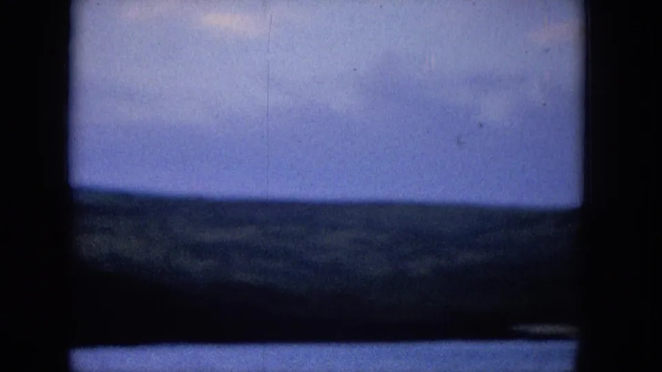 1965 archival home-movie view of a distant shoreline beneath a blue clouded sky, partially obscured by a dark vertical and horizontal frame.
