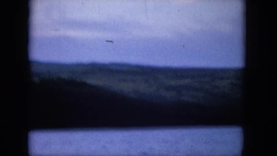 1965 archival home-movie frame showing a soft blue ocean or coastal waterline with distant land, viewed through a dark structural frame and marked by visible small-gauge film texture.