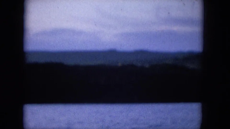 1965 archival home-movie frame showing blue sky, faint cloud cover, and dark coastal landforms behind a strong window-like grid, with grain and faded color.