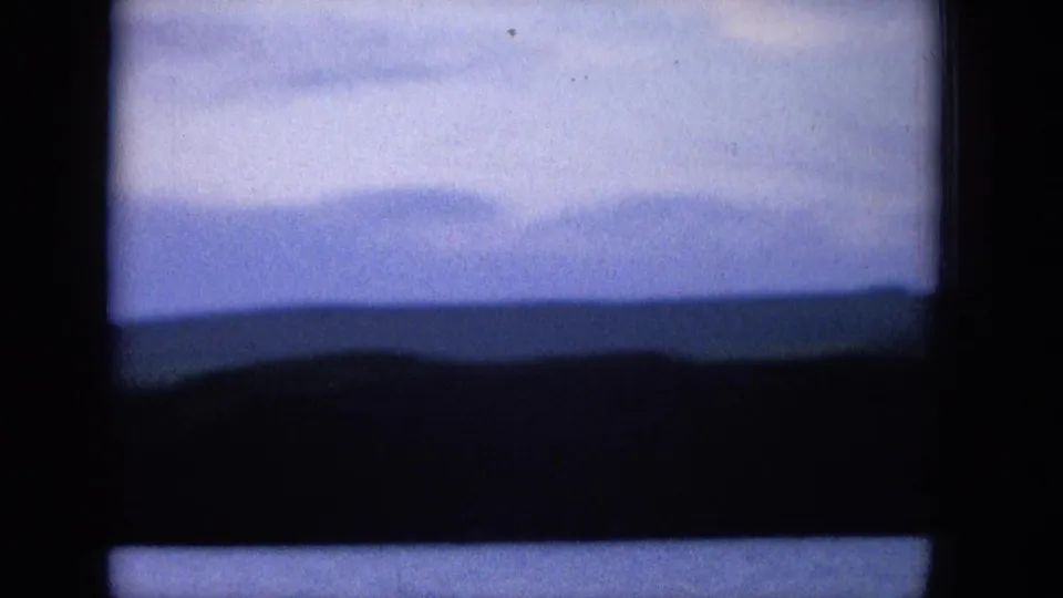 1965 8mm Hawaii landscape frame showing a soft blue sky above a dark distant sea and low hillside, seen through heavy home-movie frame edges.