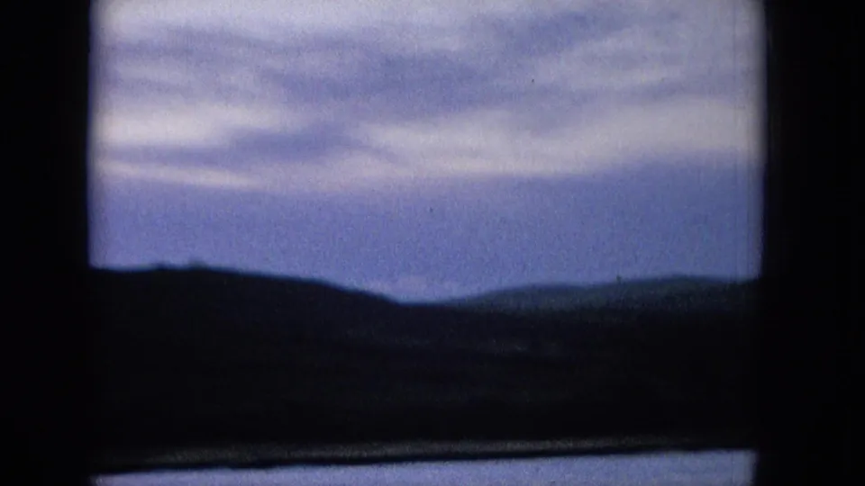 1965 home-movie frame of a Hawaiian coastal horizon, with muted clouds, blue sea tones, and dark hill forms beneath a thick frame edge.