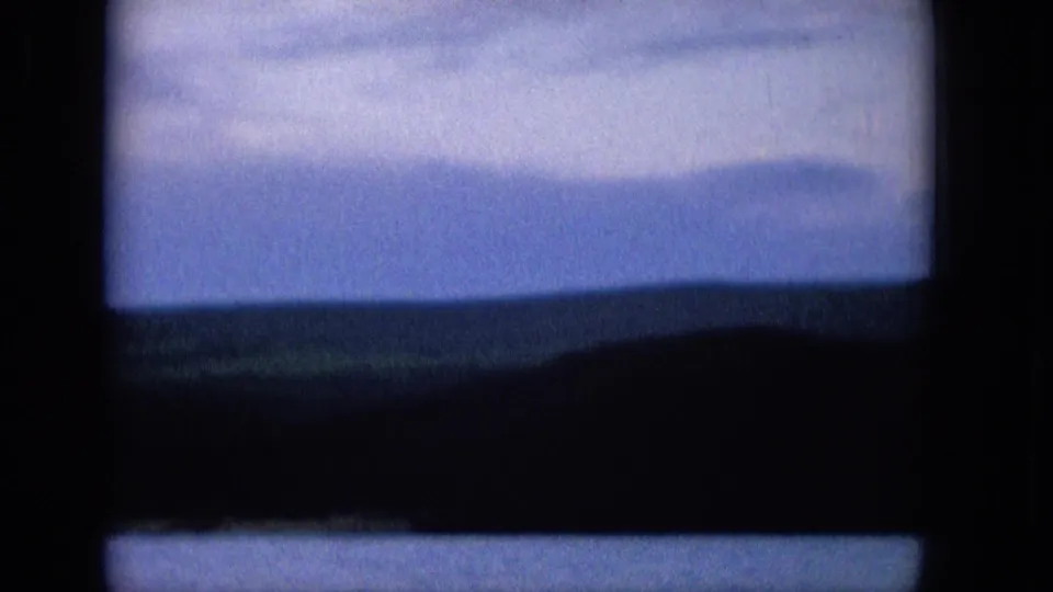 A soft 1965 8mm Hawaiian landscape frame with blue sky, distant water, and low hills partially enclosed by dark rounded film-frame edges.