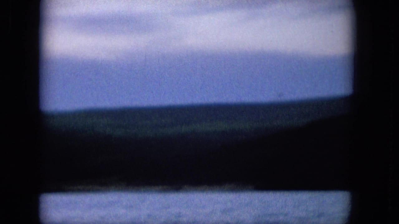 1965 8mm frame showing a coastal or lakeside view through a dark window frame, with blue water below, a distant low shoreline, and pale clouds above.