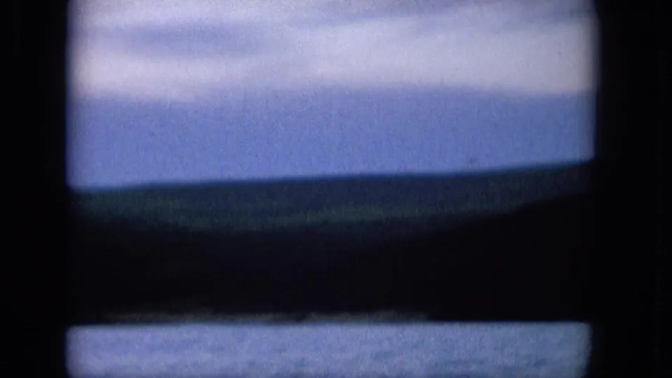 1965 8mm frame showing a coastal or lakeside view through a dark window frame, with blue water below, a distant low shoreline, and pale clouds above.