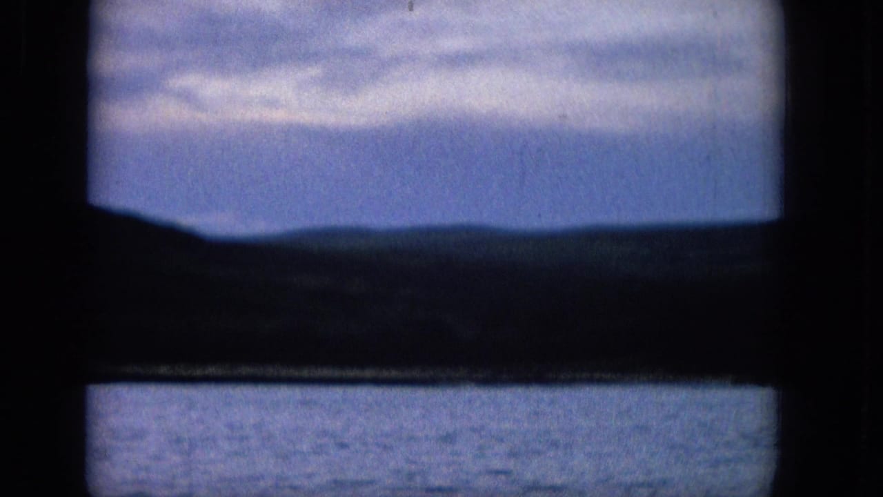 1965 8mm frame with a dark window crossbar in the foreground, calm water in the lower section, distant land across the middle, and muted blue sky with clouds.